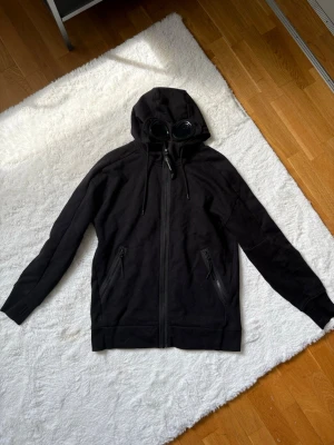Svart hoodie med glasögon från C.P. Company - Svart hoodie med dragkedja från C.P. Company. Hoodien har en unik huva med inbyggda glasögon, två fickor med dragkedja och ribbade muddar. 