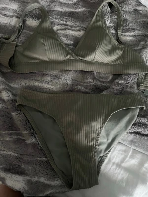 Olivgrön ribbad bikini med trekantsöverdel - Snygg olivgrön bikini med ribbat tyg och trekantsformad överdel. Både överdel och underdel har breda band och en stilren look. Materialet är stretchigt och bekvämt, perfekt för stranden eller poolen. Inga synliga loggor eller märken.