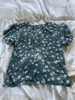 Grön blommig blus med puffärm - Söt grön blus med vita och lila blommor. Blusen har puffiga korta ärmar och rund halsringning. Tunt, luftigt material som känns perfekt för sommaren. 