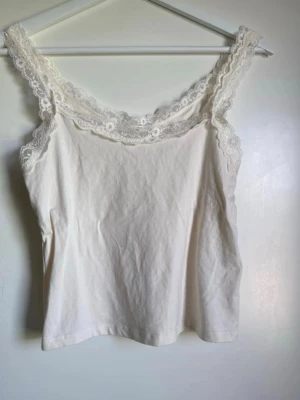 Vit spetsdetaljerad linnetopp Zara - Söt vit linnetopp från Zara med breda axelband och fina spetsdetaljer längs urringningen och axelbanden. Tillverkad i mjuk bomull, perfekt för en fräsch och stilren look.