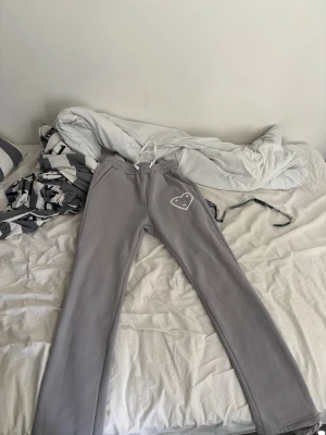 Carsicko byxor size S - Snygga grå mjukisbyxor med vitt snöre i midjan och ett broderat hjärta på ena benet. Byxorna har raka ben och resår i midjan för extra komfort. Perfekta för en chill och avslappnad stil.