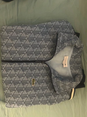 Blå mönstrad hoodie från Lacoste - Snygg blå hoodie från Lacoste med dragkedja och geometriskt mönster över hela plagget. Klassisk krokodillogga på bröstet och ribbade muddar med vita och blå ränder. Perfekt för dig som gillar streetwear och vill sticka ut lite extra.