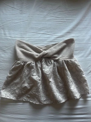 Beige broderad topp virkad - Supersöt beige topp med virkad överdel och broderade blommor i tyget. Toppen har en twistad detalj framtill och ärmar saknas. Perfekt för sommaren och ger en romantisk vibe till din outfit.