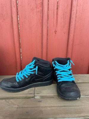 Svarta Nike sneakers med blåa skosnören - Svarta Nike sneakers i high-modell med tydlig swoosh-logga på sidan. Skorna har blåa, tjocka skosnören som sticker ut och ger en cool kontrast. Ovandelen är i läder och textil, sulan är svart och robust. Perfekta för dig som vill ha en unik streetstil.