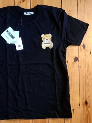 Svart Moschino t-shirt med björn - Snygg svart t-shirt från Moschino med ett broderat björnmärke på bröstet. Klassisk rund halsringning och korta ärmar. Tillverkad i mjuk bomull som känns skön mot huden. Perfekt för dig som vill ha en trendig och lekfull look.