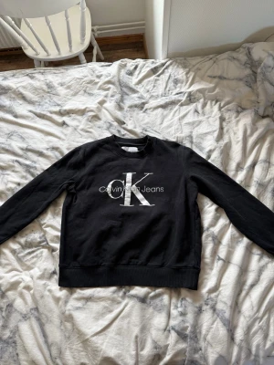 Svart sweatshirt från Calvin Klein Jeans - Svart sweatshirt från Calvin Klein Jeans med stor vit CK-logga och text på bröstet. Tröjan har rund halsringning, långa ärmar och ribbade muddar. Tillverkad i mjukt bomullsmaterial som är skönt mot huden.