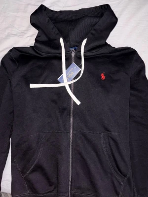 Svart hoodie från Polo Ralph Lauren - Svart hoodie med dragkedja från Polo Ralph Lauren. Klassisk design med vit dragsko, huva och röd broderad logga på bröstet. Mjuk bomullskänsla och två fickor framtill. Perfekt för en avslappnad och stilren look. Stor i storleken.