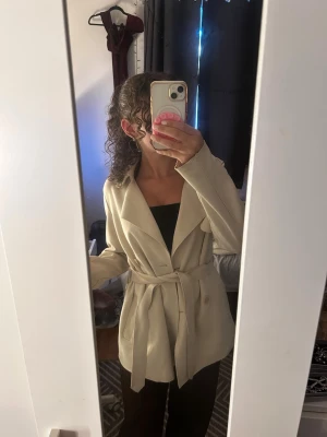 Beige trenchcoat med knytskärp - Stilren beige trenchcoat med breda slag och knytskärp i midjan. Jackan har en klassisk, rak passform och är tillverkad i ett mjukt tyg som ger en snygg och avslappnad look. Perfekt för lager-på-lager och enkel att matcha med olika outfits.