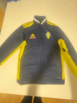 Svenska landslaget adidas jacka - Mörkblå och gul träningsjacka från adidas med Sveriges landslagsemblem och Bauhaus-logga på bröstet. Jackan har dragkedja, ståkrage och gula detaljer längs ärmar och axlar. Perfekt för fotbollsträning eller support.                                 Går för både kvinnor och män! Inga skador eller märken.