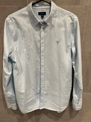 Ljusblå skjorta från Gant - Klassisk ljusblå skjorta från Gant med broderad logga på bröstet. Skjortan har lång ärm, knappar framtill och button-down krage. Perfekt för en clean och stilren look.