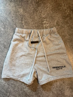 Grå shorts Essentials Fear of God - Snygga ljusgrå shorts från Essentials Fear of God med snörning i midjan och tryckt logga på benet. Mjukt material som känns skönt mot huden och perfekt för avslappnade dagar. Klassisk passform och enkel design.