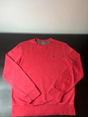 Röd crewneck från Ralph Lauren - Röd crewneck-tröja från Ralph Lauren med klassisk blå broderad logga på bröstet. Tröjan har rund halsringning, långa ärmar och ribbade muddar. Perfekt för en stilren och avslappnad look.