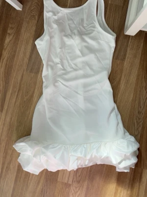 Vit baddräkt med volang - Vit baddräkt med breda axelband och rund halsringning. Baddräkten har en figurnära passform och en stor volangdetalj längst ner som ger en unik och lekfull look. Tillverkad i ett stretchigt material som känns bekvämt mot huden.