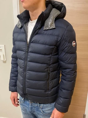 Mörkblå pufferjacka från Colmar - Snygg mörkblå pufferjacka från Colmar med hög krage och avtagbar huva. Jackan har två dragkedjeförsedda fickor framtill och Colmar-logga på ärmen. Tillverkad i ett glansigt material som ger en modern look och passar perfekt för alla årstider!