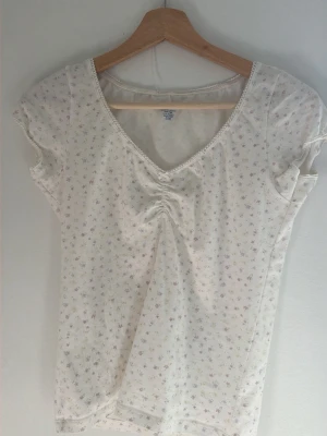 Brandy Melville - Vit topp med blommigt mönster - Söt vit topp med små blommor, v-ringning och rynkdetalj framtill. Korta ärmar och mjukt, stretchigt material. Helt ny och oanvänd. Köpte i Italien när jag var på semester. Har inte kommit till användning och säljer därav.❤️😊