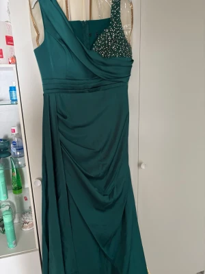 Grön långklänning med paljettdetalj - Elegant mörkgrön långklänning med asymmetrisk axel och en axel täckt av glittrande paljetter. Klänningen har draperad front och slät baksida, vilket ger en snygg och stilren siluett. Perfekt för speciella tillfällen när du vill sticka ut. Priset går att diskutera, den är köpt för 3000kr💗