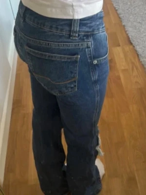 Blå bootcut jeans med hål - Säljer ett par blå bootcut jeans med hål och slitningar över knäna och råa kanter. Jeansen har klassisk femficksdesign, låg midja och är tillverkade i denim. Perfekta för en avslappnad och trendig look.
