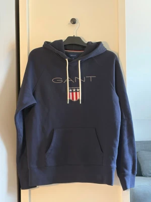 Blå hoodie från GANT med logga - Mörkblå hoodie från GANT med broderad logga och sköld på bröstet. Klassisk känguruficka framtill, vita dragsnören i huvan och ribbade muddar. Perfekt för en avslappnad och stilren look.