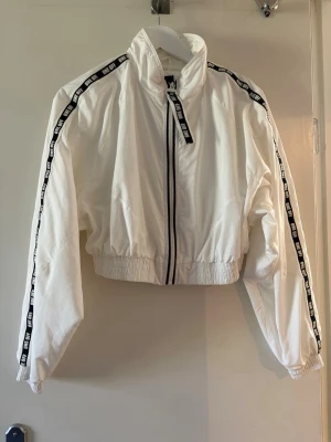 Vit tunnjacka med svarta detaljer - Snygg vit jacka med svarta band längs ärmarna där det står 'ONE OFF'. Jackan har hög krage, dragkedja framtill och resår i midja och ärmslut. Croppad modell som ger en sportig vibe. Perfekt till streetwear-looken.