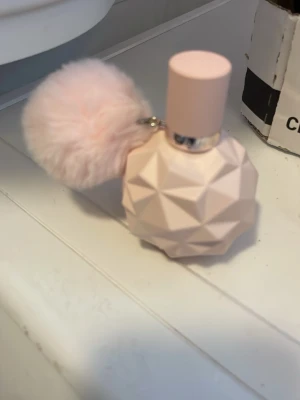 Ariana Grande Sweet Like Candy parfym - Säljer en Ariana Grande Sweet Like Candy parfym i en ljusrosa, geometrisk flaska med en fluffig rosa boll som dekoration. Flaskan har en matt finish och ett silverfärgat band runt halsen där det står 'Ariana Grande'. Perfekt som present eller till dig själv!