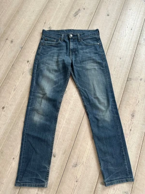 Blå raka jeans från Levi's modell 511 - Snygga blå jeans från Levi's med klassisk rak passform och fem fickor. Jeansen har en  tvättad look med slitningar på låren och är tillverkade i bomull. Har ett litet hål på vänster lår. Storlek 29x30. 
