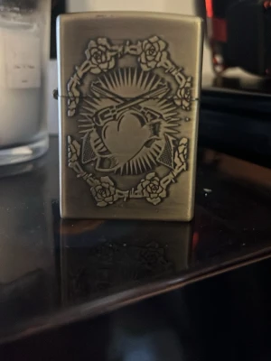 Zippo tändare med hjärtmotiv - Metallisk Zippo tändare i mässingsfärg med graverat hjärta, rosor och strålande solstrålar. Klassisk rektangulär form och flip-top lock. Perfekt för dig som gillar unika accessoarer med detaljerad design.