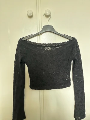 Svart spetstopp - Snygg svart croppad topp i spets med lång ärm och v-ringad hals. Toppen har ett transparent spetsmönster och sitter offshoulder.