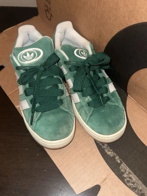 Adidas Campus gröna sneakers - Gröna Adidas Campus sneakers i mocka med vita klassiska ränder på sidorna och vit sula. Skorna har gröna skosnören och Adidas logga på plösen. Snygg retrostil som passar perfekt till streetwear.