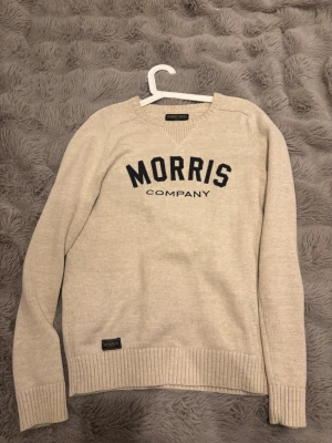 Beige tröja från Morris Gents - Snygg beige stickad tröja från Morris Gents med svart text 'MORRIS COMPANY' på bröstet. Tröjan har rund halsringning, ribbade muddar och nederkant samt en liten patch nertill. Perfekt för en stilren och avslappnad look.