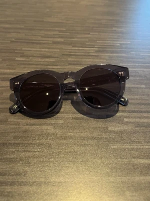 Chimi solglasögon - Snygga svarta solglasögon med rund form och transparent båge. Glasen är mörka och bågarna har en stilren design som passar till de flesta outfits. Perfekta för soliga dagar och ger en cool vibe.