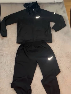 Svart Nike träningsset med huva - Snyggt svart träningsset från Nike med jacka och byxor. Jackan har huva, dragkedja och fickor, samt reflekterande Nike-logga på bröstet och ärmen. Byxorna har resår i midjan och reflekterande logga på benet. Perfekt för löpning eller gym. Materialet är lätt och snabbtorkande. Storlek M
