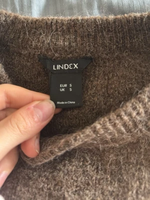 Brun stickad tröja från Lindex - Mysig brun stickad tröja från Lindex i storlek S. Tröjan har en klassisk rund halsringning och är tillverkad i ett fluffigt, mjukt material som passar perfekt för kyliga dagar. Enkel och stilren design som är lätt att matcha med olika outfits.
