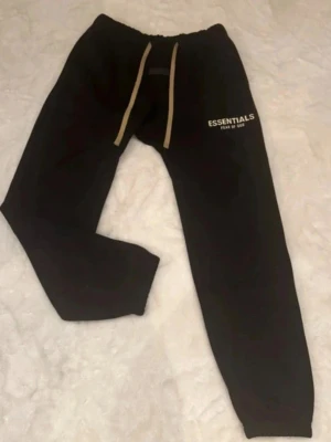 Svarta Essentials mjukisbyxor - Säljer ett par svarta mjukisbyxor från Fear of God Essentials med vit logga på benet och beige dragsko i midjan. Byxorna har resår i midjan och vid bensluten, samt en avslappnad passform i mjukt material.