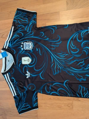Argentina Adidas fotbollströja svart/blå. Storlek .S.M - Helt ny  svart fotbollströja från Adidas med Argentinas landslagslogga och FIFA-märke på bröstet. Tröjan har ett unikt blått mönster över hela tyget och klassiska vita Adidas-ränder på axlarna. Perfekt för dig som älskar fotboll och vill sticka ut på planen.