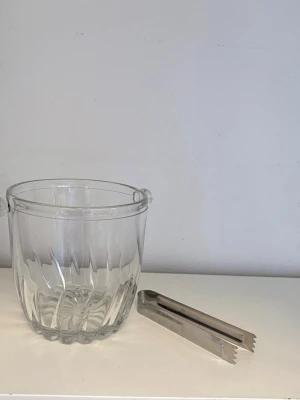 Isbehållare i glas med tång - Säljer en stilren isbehållare i klart glas med räfflad design och två små handtag på sidorna. Medföljer en silvrig istång i metall. Perfekt till drinkbordet eller festen för att hålla isen kall och servera smidigt.