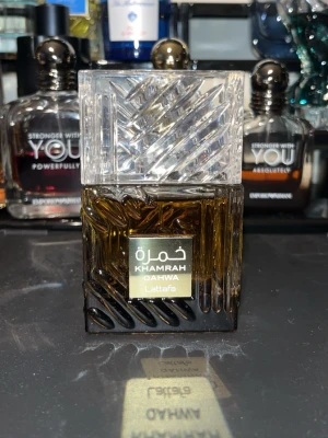 Lattafa Khamrah Qahwa parfym - Lattafa Khamrah Qahwa är en parfym i en elegant, fyrkantig flaska med mörkbrun glasbotten och ett genomskinligt, mönstrat lock. Flaskan har en lyxig känsla och etiketten är guld- och vitfärgad med arabiska och engelska texter. 98/100ml kvar ungefär