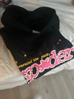 Svart hoodie från SP5DER med tryck - Svart hoodie från SP5DER med färgglatt tryck framtill där det står 'Nevermind the punk, SP5DER' i gult och rosa med vita prickar. Hoodien har huva och en avslappnad streetwear-stil, perfekt för dig som gillar unika plagg. Äkta 