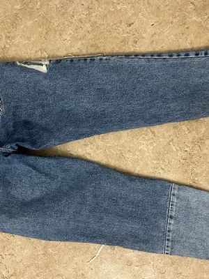 Blå raka jeans från Zara - Säljer ett par blå raka jeans från Zara med slitningar vid fickan och en ljusare panel längst ner på ena benet. Klassisk femficksmodell i denim med råa kanter och trendig vintagekänsla. Perfekta för dig som gillar en avslappnad och cool stil.