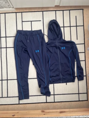 Under Armour mörkblå träningsset - Snyggt träningsset från Under Armour i mörkblått med ljusblå detaljer. Setet består av en zip-hoodie med huva och matchande byxor med resår i midjan. Båda plaggen har Under Armour-logga och är tillverkade i ett lätt och stretchigt material, perfekt för träning.