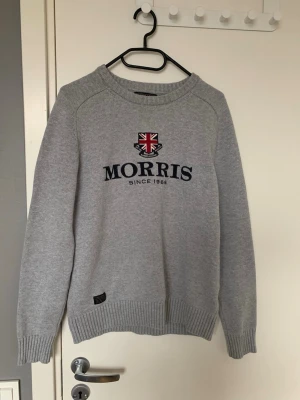 Grå stickad tröja från Morris - Snygg grå stickad tröja från Morris med broderad logga och brittisk flagga framtill. Tröjan har rund hals, långa ärmar och ribbade muddar vid ärmslut och nederkant. Perfekt för en stilren och klassisk look. Den är skön och väldigt fint material. 