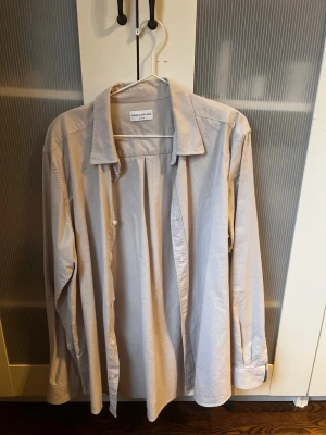 Ljusgrå skjorta från These Glory Days - Stilren ljusgrå skjorta från These Glory Days i bomull. Klassisk krage, knäppning framtill och långa ärmar med manschett. Perfekt för en clean och enkel look.
