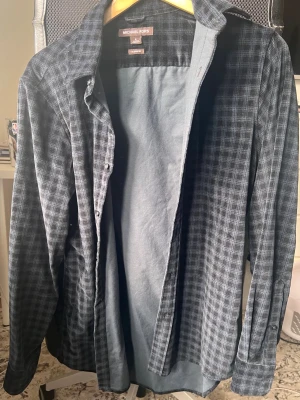 Rutig skjorta från Michael Kors - Snygg rutig skjorta från Michael Kors i mörkblå och grå toner. Skjortan har klassisk krage, långa ärmar och knäppning framtill. Perfekt för dig som gillar en stilren och avslappnad look.