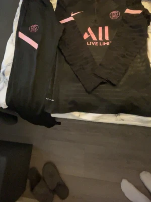 Svart PSG träningsset från Nike - Säljer ett svart PSG träningsset från Nike med rosa detaljer. Setet består av långärmad tröja och byxor, båda med PSG-logga och Nike Swoosh. Tröjan har trycket 'ALL LIVE LIMITLESS' i rosa framtill. Perfekt för fotbollsträning eller chill.