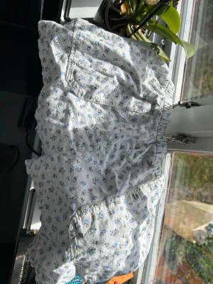 Vita blommiga shorts med resårmidja - Säljer ett par vita shorts med små blå blommor och resår i midjan. Shortsen har en avslappnad passform och är perfekta för varma dagar. De har fickor på sidorna och är gjorda i ett mjukt bomullstyg som känns skönt mot huden.