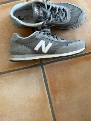 Grå New Balance 515 sneakers - Snygga grå sneakers från New Balance, modell 515. Skorna har mesh- och mockadetaljer, klassisk vit N-logga på sidan och vit sula med svart yttersula. Perfekta för en stilren och sportig look.