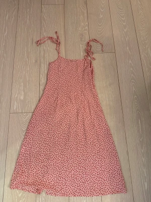 Rosa klänning med hjärtmönster - Söt rosa klänning med små röda hjärtan och knytbara axelband. Klänningen har en lätt utsvängd form och är ärmlös, perfekt för varma dagar. Tillverkad i ett mjukt och luftigt material som känns bekvämt mot huden.