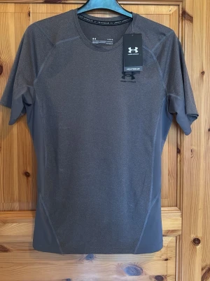 Under Armour Compression Shirt - Snygg grå kortärmad träningströja från Under Armour med HeatGear-teknologi. Tröjan är tillverkad i ett lätt och stretchigt funktionsmaterial som andas och transporterar bort fukt. Perfekt för intensiva träningspass eller löpning. Helt ny och oanvänd. Kvitto finns. Säljer pga att jag inte gymmar. Pris kan alltid diskuteras💫💫