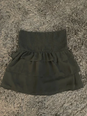 Svart volangkjol från BikBok - Supersöt svart kortkjol från BikBok med bred smock i midjan och dubbla volanglager. Kjolen har en flowy känsla och passar perfekt till en trendig look. Enkel att matcha med olika toppar för en cool outfit.