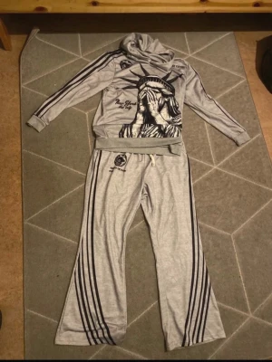 Grått loungewear-set med tryck - Grått loungewear-set med hoodie och byxor. Setet har svarta ränder längs ärmar och ben, samt tryck med Frihetsgudinnan och texten 'New York' och 'Los Angeles'. Byxorna har dragsko i midjan och vida ben. Perfekt för en avslappnad stil hemma.