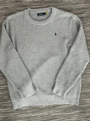 Grå stickad tröja från Polo Ralph Lauren - Snygg grå stickad tröja från Polo Ralph Lauren med klassiskt rund halsringning och diskret blå logga broderad på bröstet. Tröjan har en strukturerad stickning och ribbade muddar vid ärmslut och nederkant. Perfekt för lager-på-lager-stil. Storlek L men sitter som XL